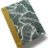 This handcrafted fabric journal features a bold jungle leaf pattern in deep green and cream, inspired by tropical foliage. The gold book cloth spine adds a sophisticated contrast, making it a stylish and durable choice for journaling, note-taking, or sketching. Perfect for writers, artists, and stationery enthusiasts, this unique fabric-covered notebook combines elegance with functionality.