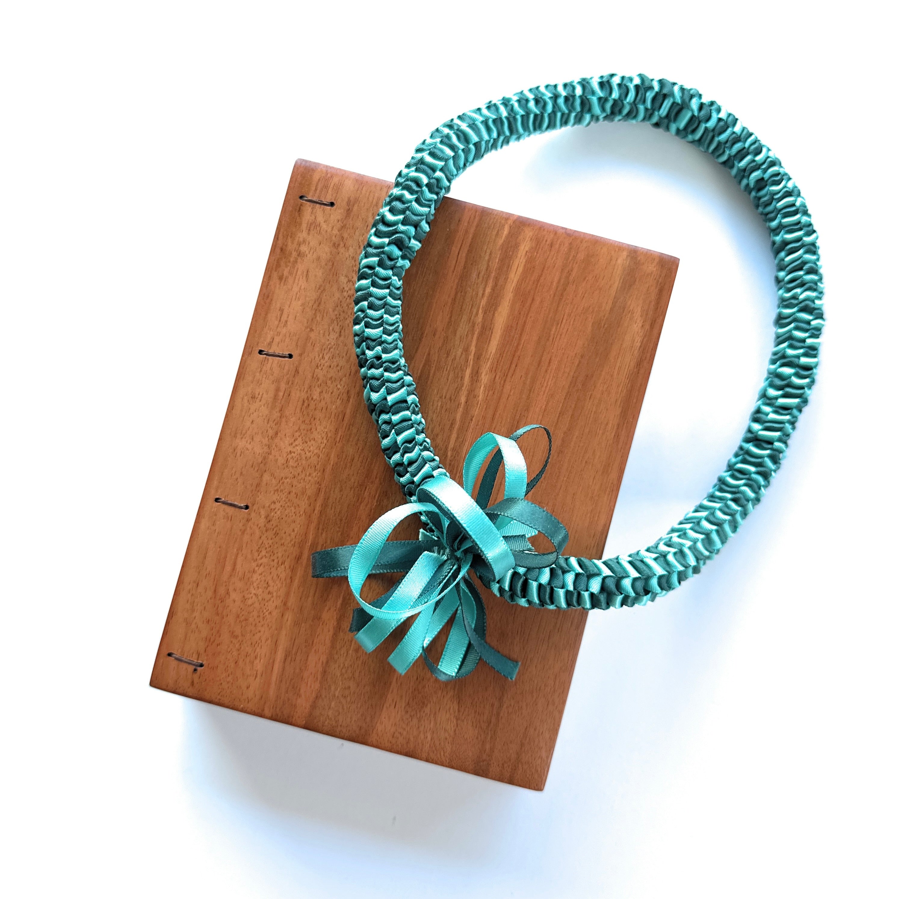 Handmade Lychee wood Coptic journal paired with a teal Hawaiian ribbon lei. The wooden cover has a smooth, natural grain, and the journal lays flat when open.