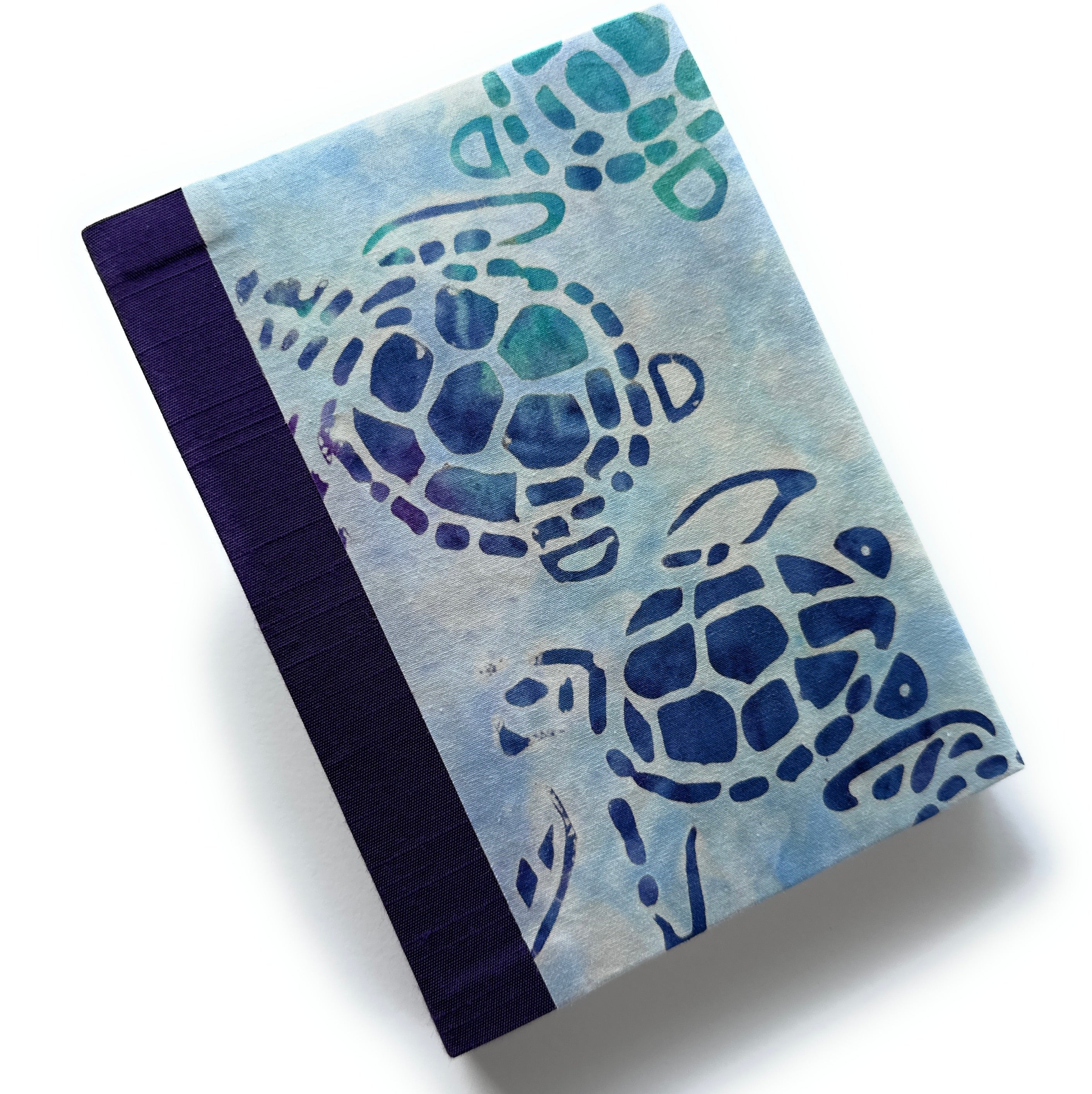 A handmade journal featuring a batik-style cover with blue and green sea turtles (“honu”) swimming across a soft watercolor background, paired with a deep indigo spine.