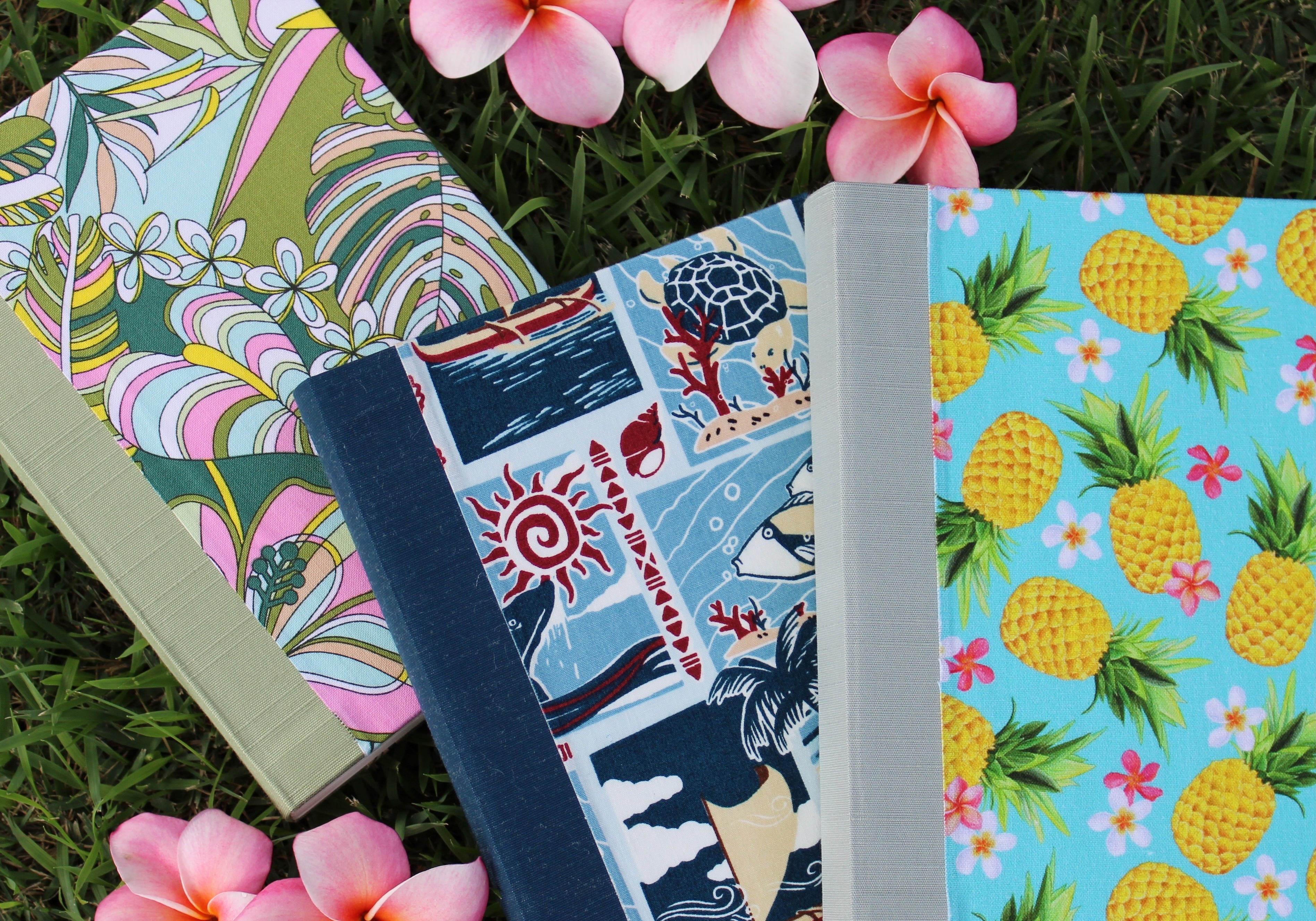 Three colorful journals with vibrant Hawaiian-themed covers are arranged on green grass, surrounded by pink and white plumeria flowers. The first journal features a tropical leaf and flower pattern, the second has a design with sea turtles, fish, and traditional Hawaiian motifs, and the third showcases bright yellow pineapples with pink flowers on a blue background. The image evokes a sense of the islands’ natural beauty and the artistic spirit of the journals.