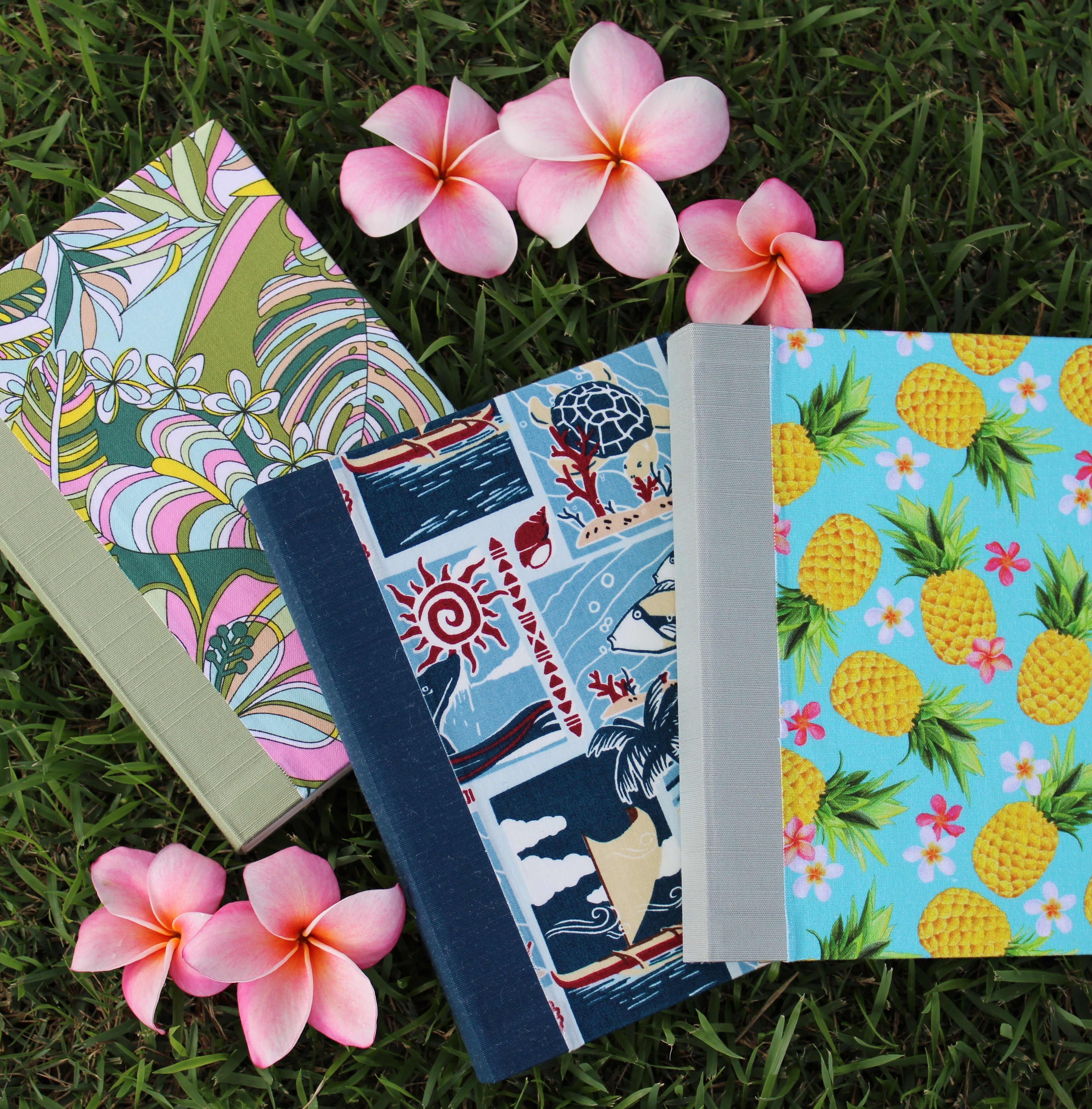 Three colorful journals with vibrant Hawaiian-themed covers are arranged on green grass, surrounded by pink and white plumeria flowers. The first journal features a tropical leaf and flower pattern, the second has a design with sea turtles, fish, and traditional Hawaiian motifs, and the third showcases bright yellow pineapples with pink flowers on a blue background. The image evokes a sense of the islands’ natural beauty and the artistic spirit of the journals.