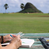 A person is sketching outdoors at a rustic wooden table, with a scenic view of a grassy field with the distinctive, Mokoliʻi island in the background. The individual holds a pencil in one hand and several other pencils in the other. A box of art supplies, including colored pencils, is positioned on the table. The setting suggests a peaceful and inspiring environment for creative work, with a clear sky and palm trees adding to the tranquil ambiance.