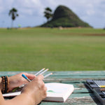 A person is sketching outdoors at a rustic wooden table, with a scenic view of a grassy field with the distinctive, Mokoliʻi island in the background. The individual holds a pencil in one hand and several other pencils in the other. A box of art supplies, including colored pencils, is positioned on the table. The setting suggests a peaceful and inspiring environment for creative work, with a clear sky and palm trees adding to the tranquil ambiance.