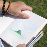 A person is holding an open sketchbook featuring a colored pencil drawing of Mokoliʻi, a distinctive island in Hawaii. The drawing captures the island’s recognizable shape and the surrounding ocean under a cloudy sky. The individual has a few pencils in one hand and wears beaded bracelets. The grassy background and the person’s relaxed posture suggest a peaceful outdoor setting, ideal for artistic inspiration and sketching.