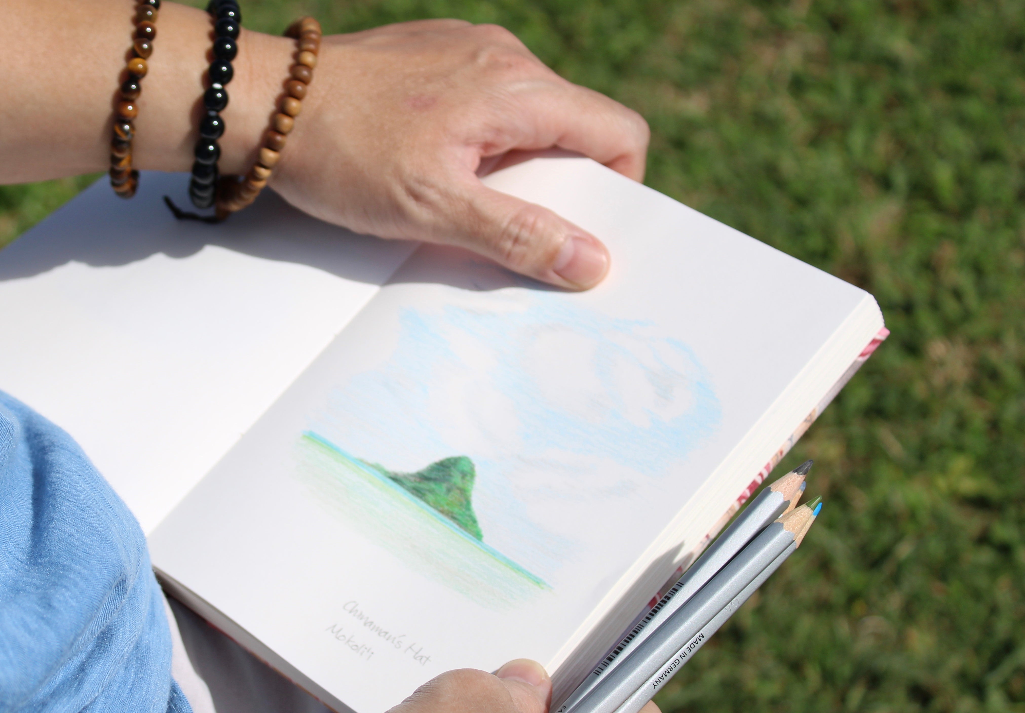 A person is holding an open sketchbook featuring a colored pencil drawing of Mokoliʻi, a distinctive island in Hawaii. The drawing captures the island’s recognizable shape and the surrounding ocean under a cloudy sky. The individual has a few pencils in one hand and wears beaded bracelets. The grassy background and the person’s relaxed posture suggest a peaceful outdoor setting, ideal for artistic inspiration and sketching.