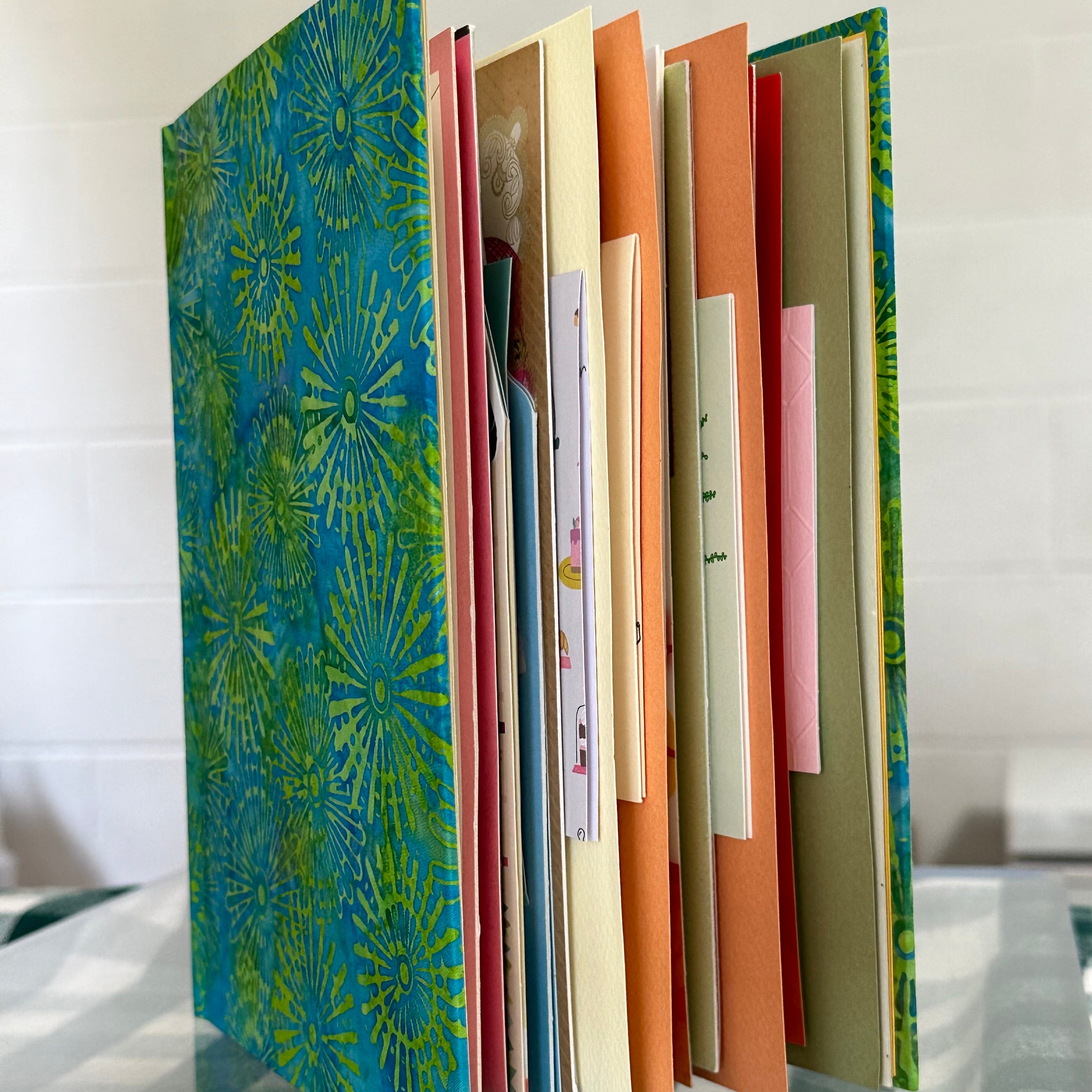 A custom-made book stands upright on a clear glass table, displaying its colorful pages and covers. The cover features a vibrant blue and green pattern reminiscent of fireworks or sunbursts. The pages inside include a variety of greeting cards, each with different colors and designs, creating a visually appealing and unique collage. The image highlights the personalized and creative nature of the book, showcasing its potential as a keepsake or gift.