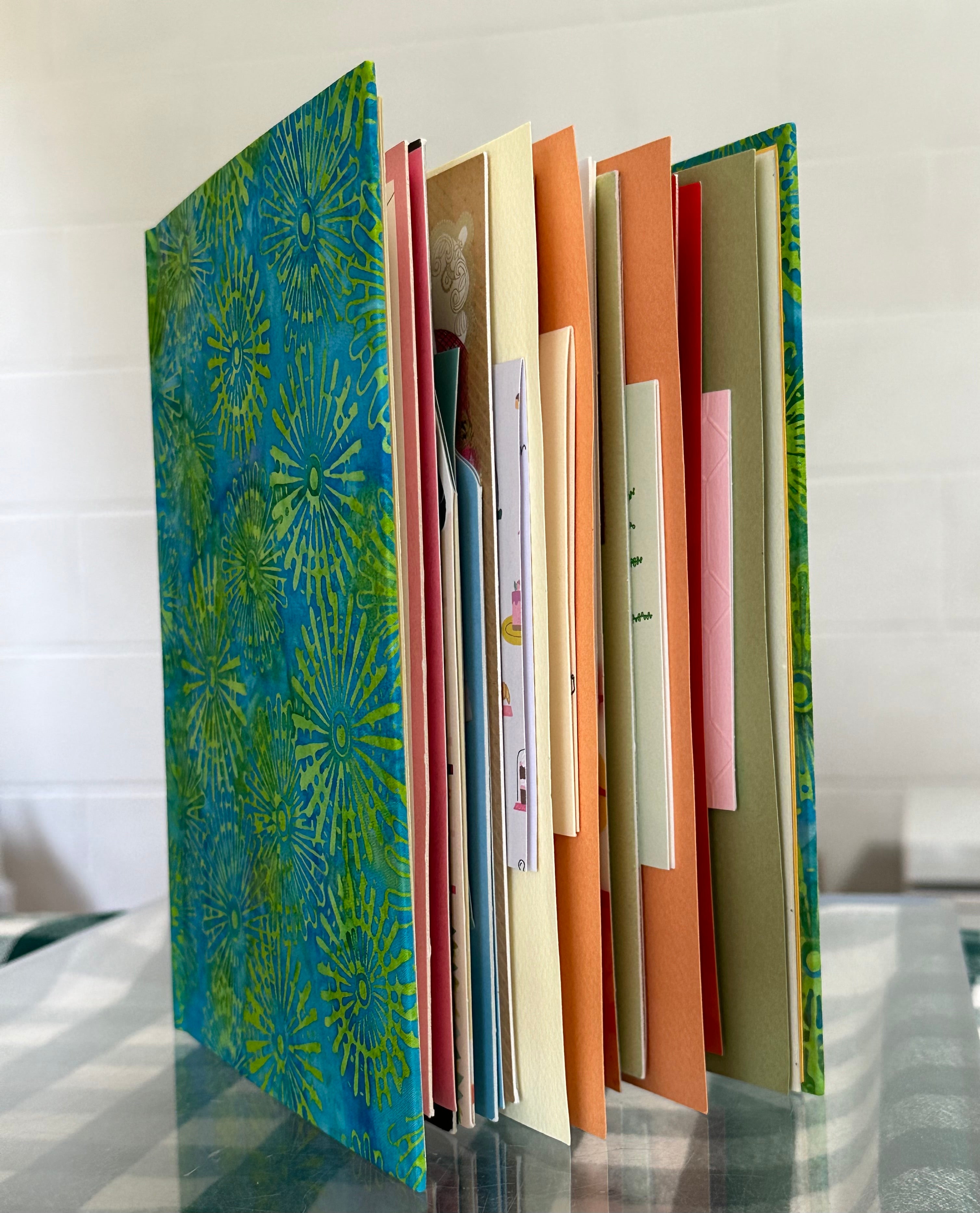 A custom-made book stands upright on a clear glass table, displaying its colorful pages and covers. The cover features a vibrant blue and green pattern reminiscent of fireworks or sunbursts. The pages inside include a variety of greeting cards, each with different colors and designs, creating a visually appealing and unique collage. The image highlights the personalized and creative nature of the book, showcasing its potential as a keepsake or gift.