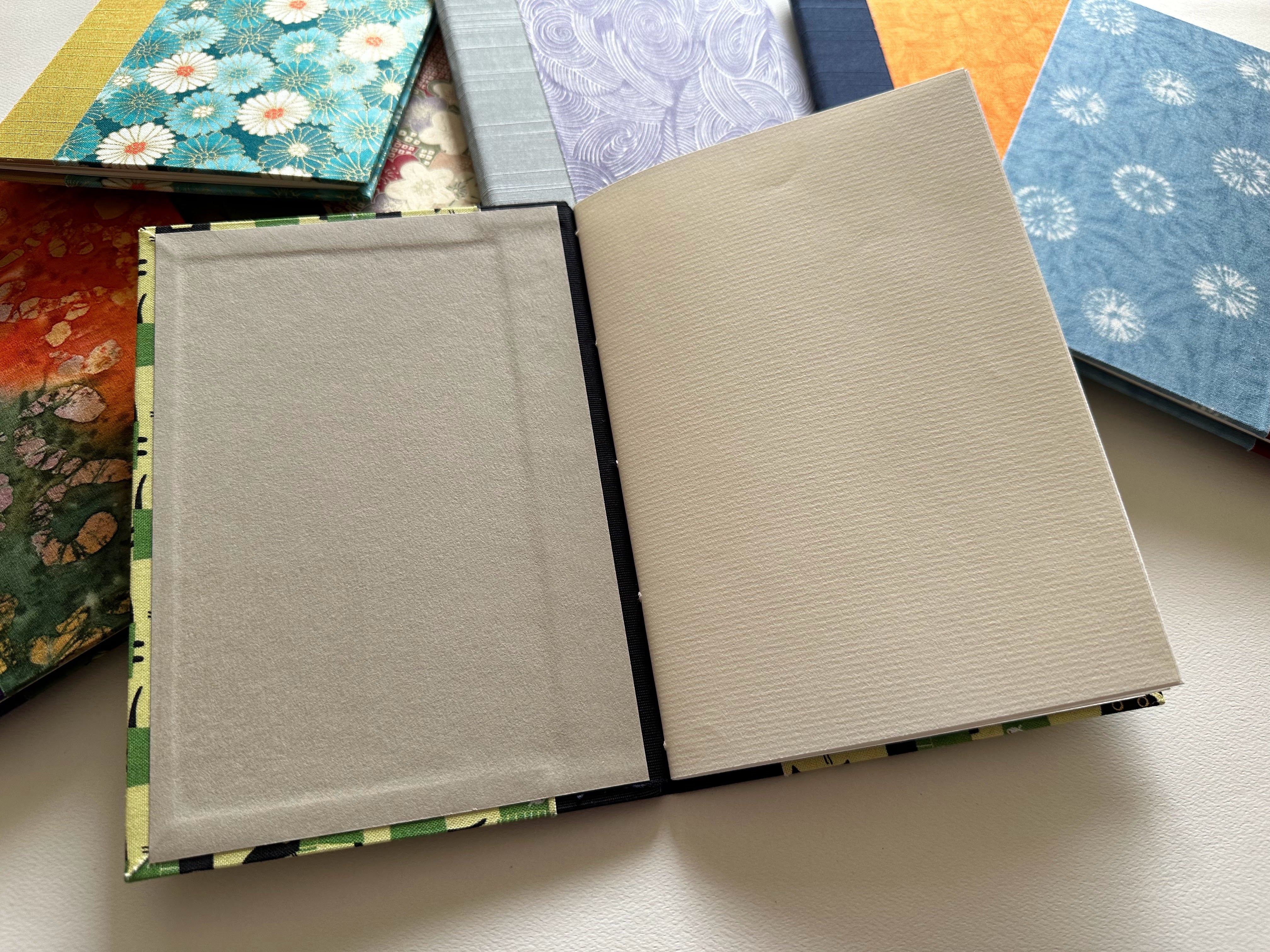An open handmade keepsake card displaying high-quality textured pages and a fabric-covered hardcover. Perfect for personalized messages, well-wishes, or special occasions, this booklet provides ample space for writing while ensuring a beautifully crafted presentation. Surrounded by other unique handcrafted designs.
