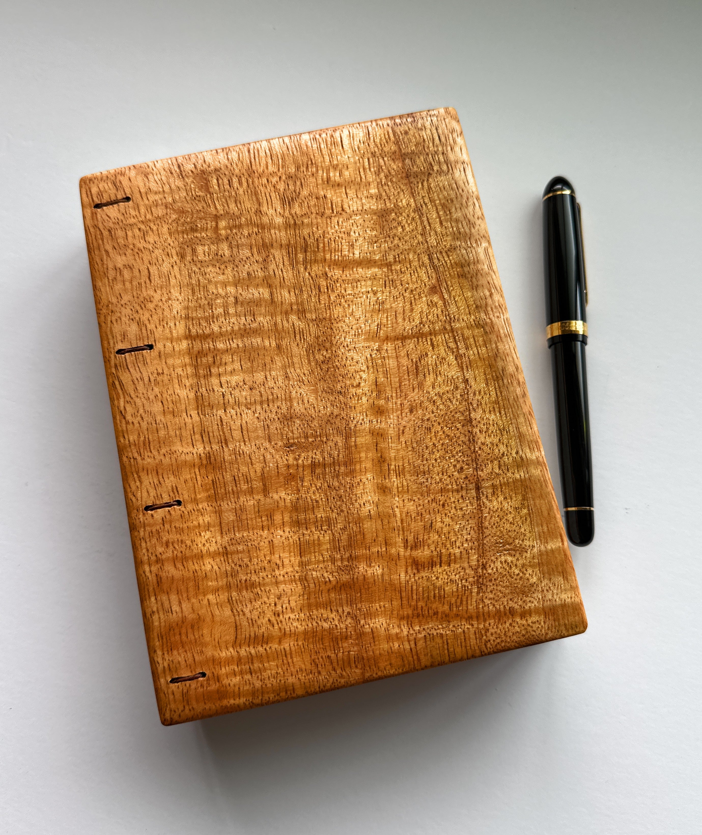 A beautifully handcrafted Mango Wood Coptic Journal with rich natural grain, placed next to a black and gold fountain pen for scale. This artisanal wooden notebook is ideal for journaling, sketching, or creative writing, blending timeless craftsmanship with functionality.