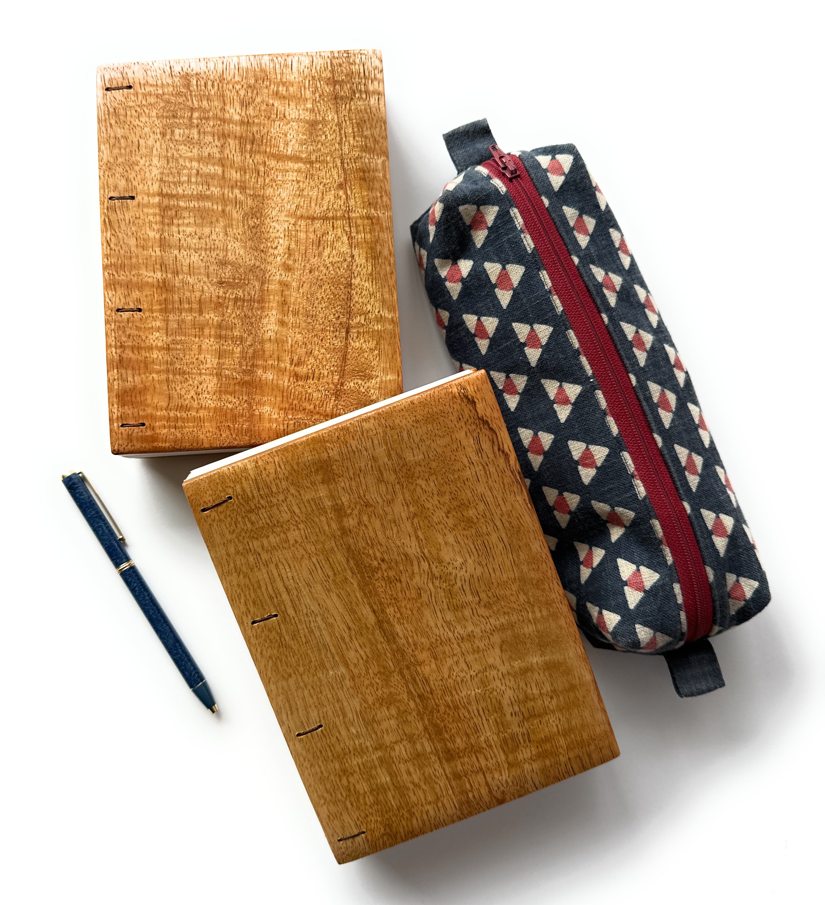 Two handcrafted Mango Wood Coptic Journals displayed alongside a patterned fabric pencil case and a navy blue fountain pen. These artisanal wooden notebooks feature exposed spines for flexibility and durability, making them perfect for journaling, sketching, or note-taking. A stylish and functional set for writers, artists, and creatives who appreciate craftsmanship and natural materials.