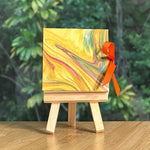 Marble-patterned miniature book on a wooden easel with an orange ribbon against a blurred green foliage background