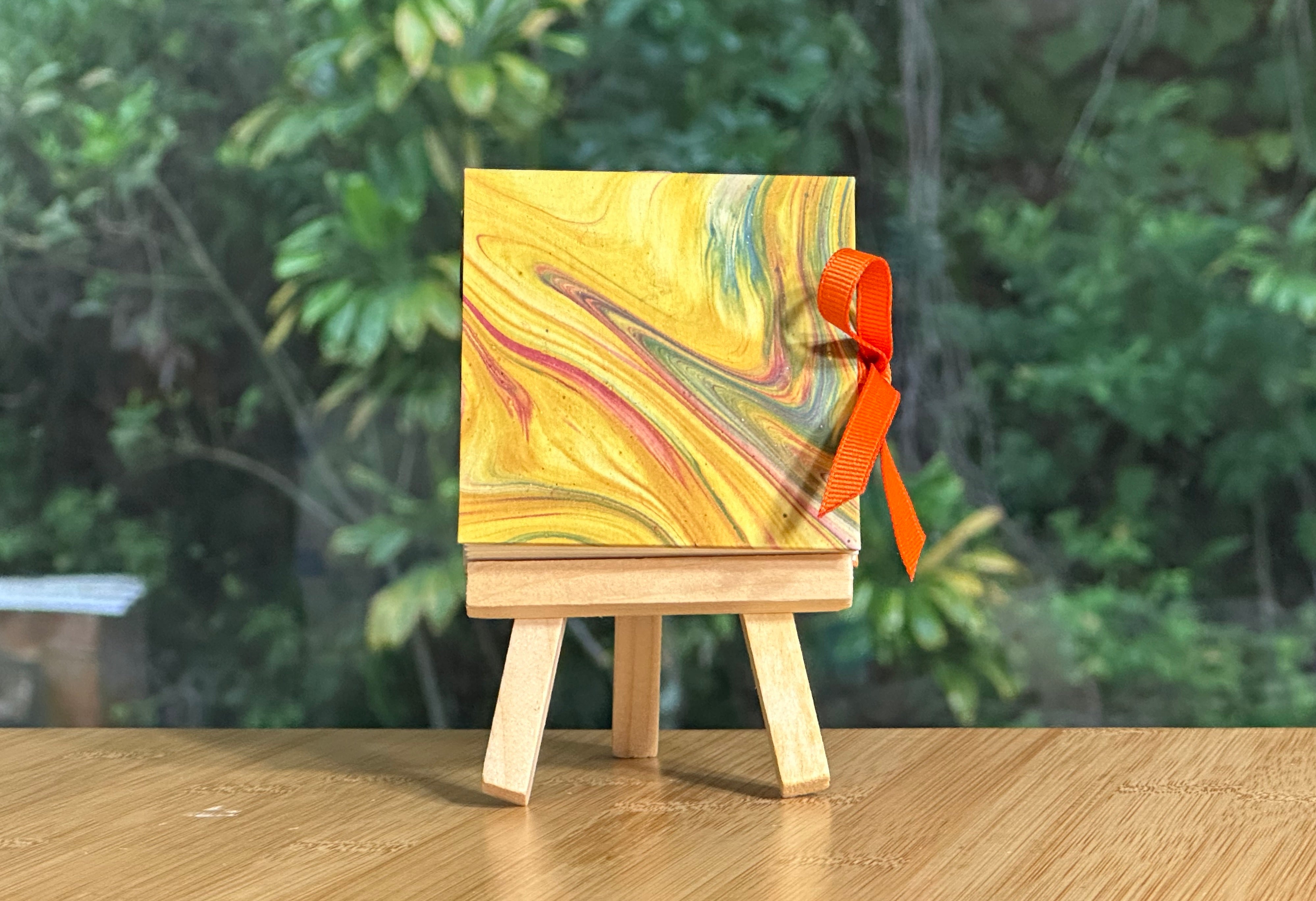 Marble-patterned miniature book on a wooden easel with an orange ribbon against a blurred green foliage background