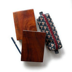 Two handcrafted Koa wood Coptic journals showcasing their rich wood grain and polished finish. A fabric pencil case with a geometric pattern and a red zipper is placed beside them, along with a navy blue and gold pen.