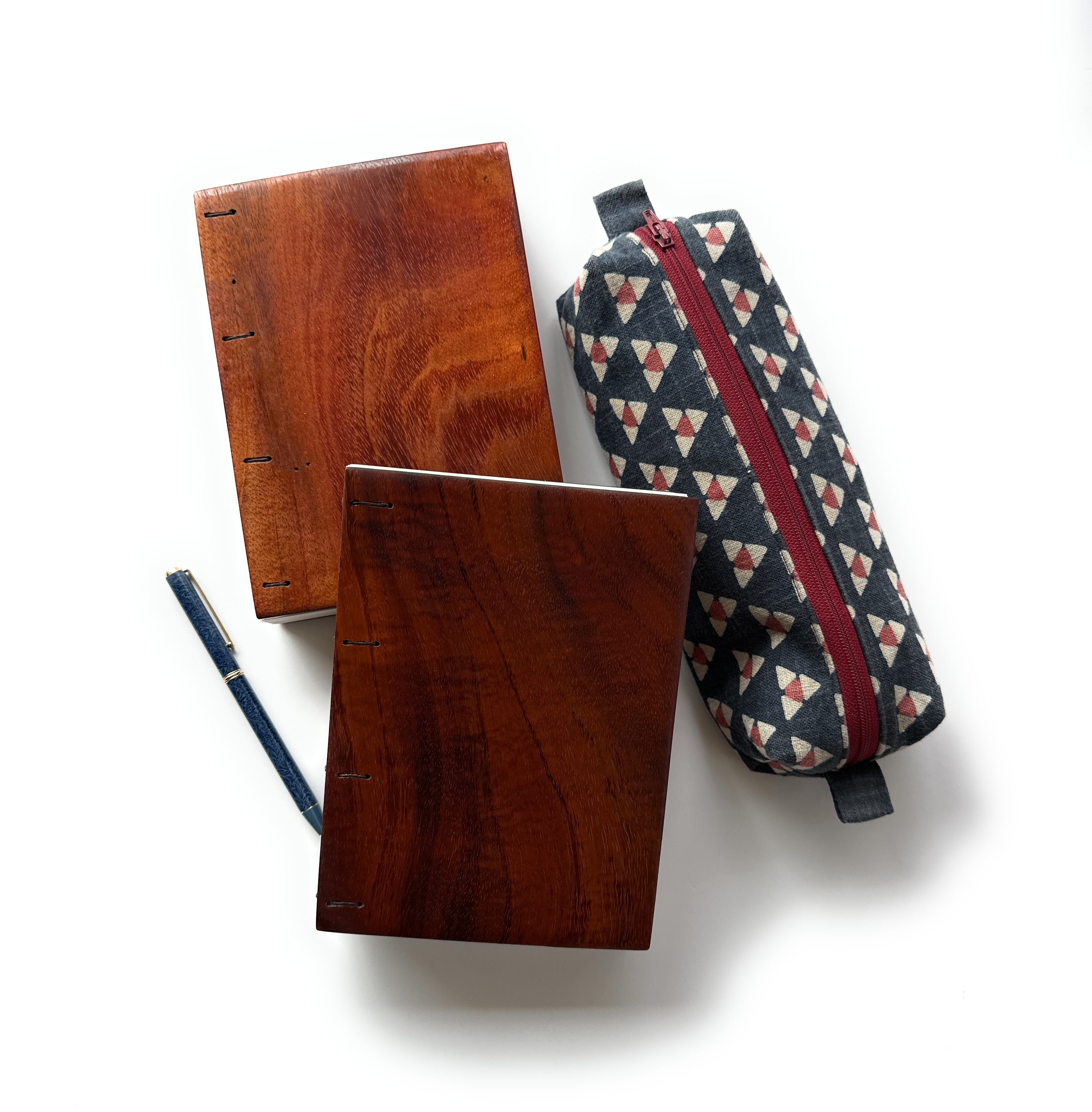 Two handcrafted Koa wood Coptic journals showcasing their rich wood grain and polished finish. A fabric pencil case with a geometric pattern and a red zipper is placed beside them, along with a navy blue and gold pen.