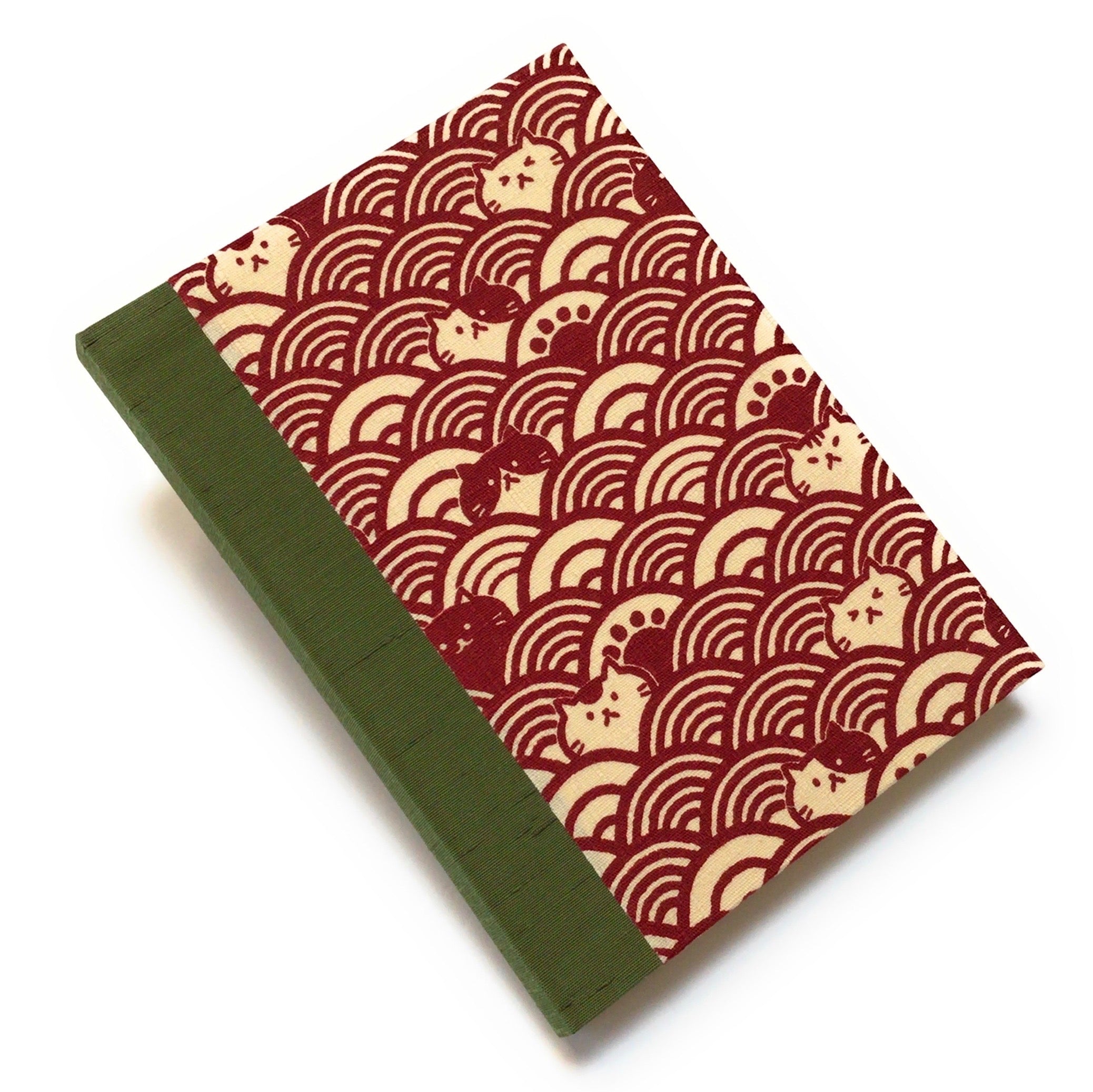 This handcrafted cat journal features a unique red and beige fabric cover with a Japanese wave pattern intertwined with whimsical feline faces. The sage green  bookbinding cloth covers the spine and is Ideal for cat lovers and stationery enthusiasts, this artisan notebook adds charm to any collection.