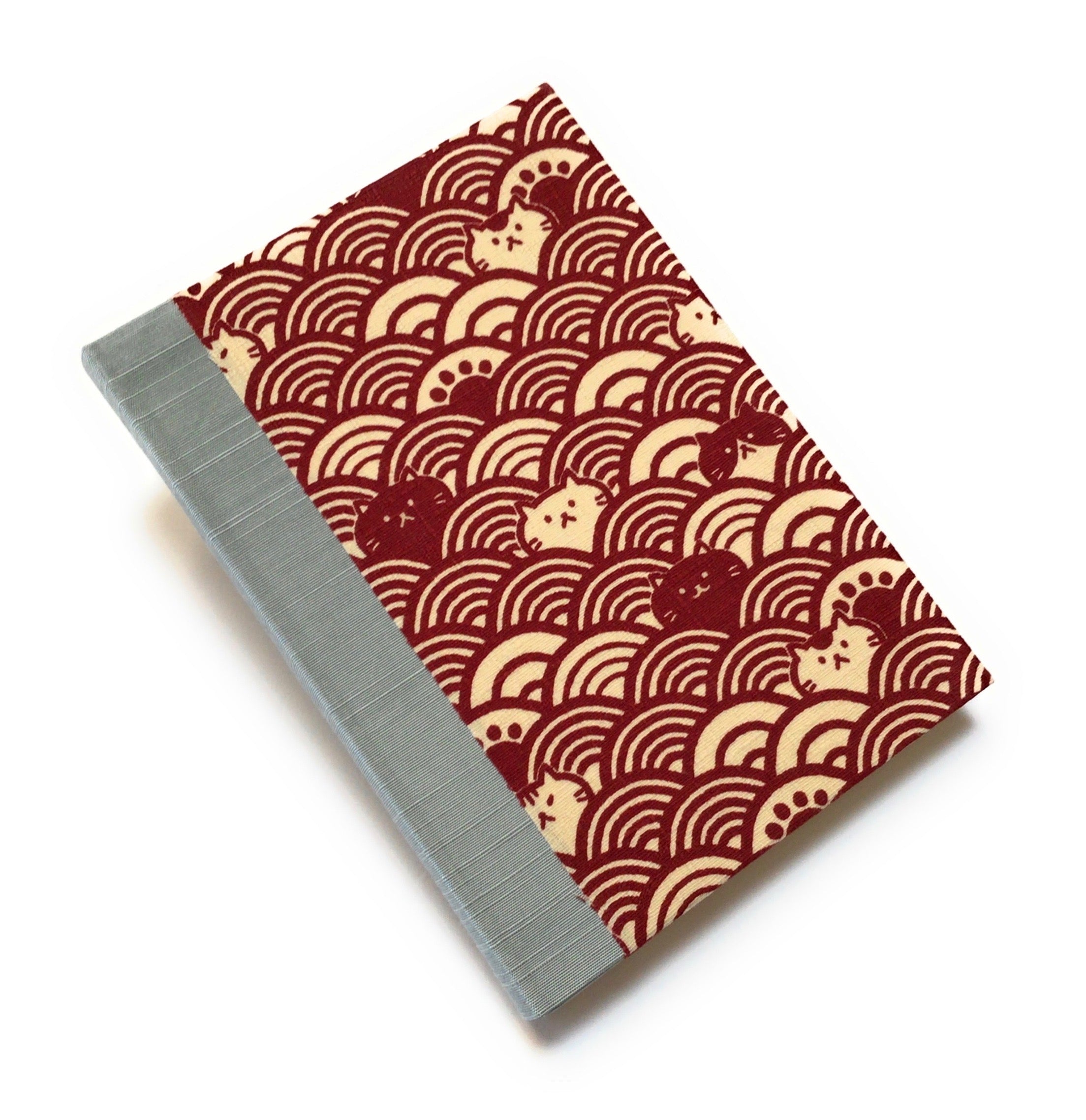 This handcrafted cat journal features a unique red and beige fabric cover with a Japanese wave pattern intertwined with whimsical feline faces. The blue gray bookbinding cloth covers the spine and is Ideal for cat lovers and stationery enthusiasts, this artisan notebook adds charm to any collection.