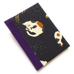 This handmade journal features a black fabric cover adorned with a serene sleeping calico cat design. Bound with a sturdy purple bookbinding cloth, this notebook is perfect for journaling, sketching, or note-taking.