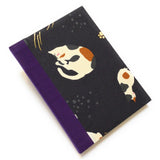 This handmade journal features a black fabric cover adorned with a serene sleeping calico cat design. Bound with a sturdy purple bookbinding cloth, this notebook is perfect for journaling, sketching, or note-taking.