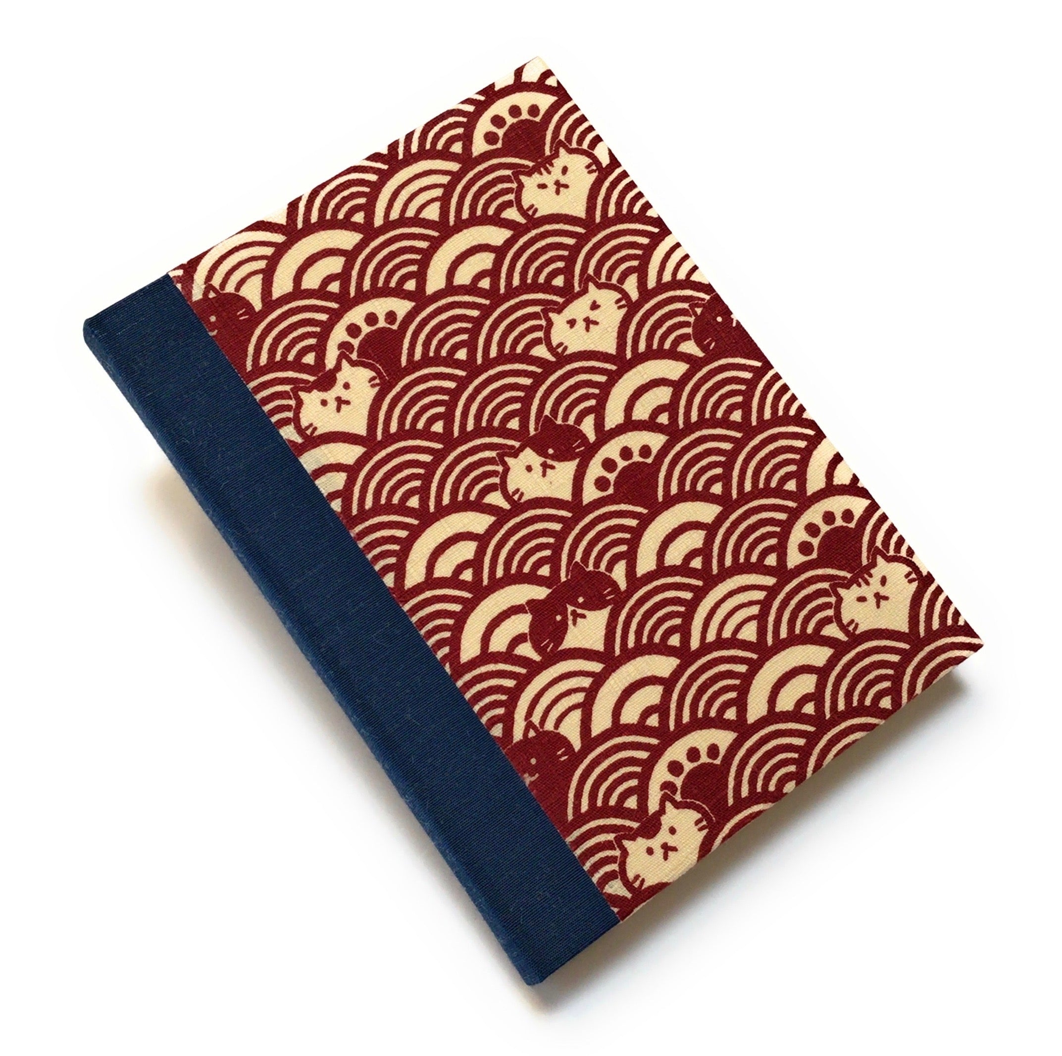 This handcrafted cat journal features a unique red and beige fabric cover with a Japanese wave pattern intertwined with whimsical feline faces. The dark blue  bookbinding cloth covers the spine and is Ideal for cat lovers and stationery enthusiasts, this artisan notebook adds charm to any collection.