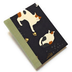 This handmade journal features a black fabric cover adorned with a serene sleeping calico cat design. Bound with a sturdy sage green bookbinding cloth, this notebook is perfect for journaling, sketching, or note-taking.