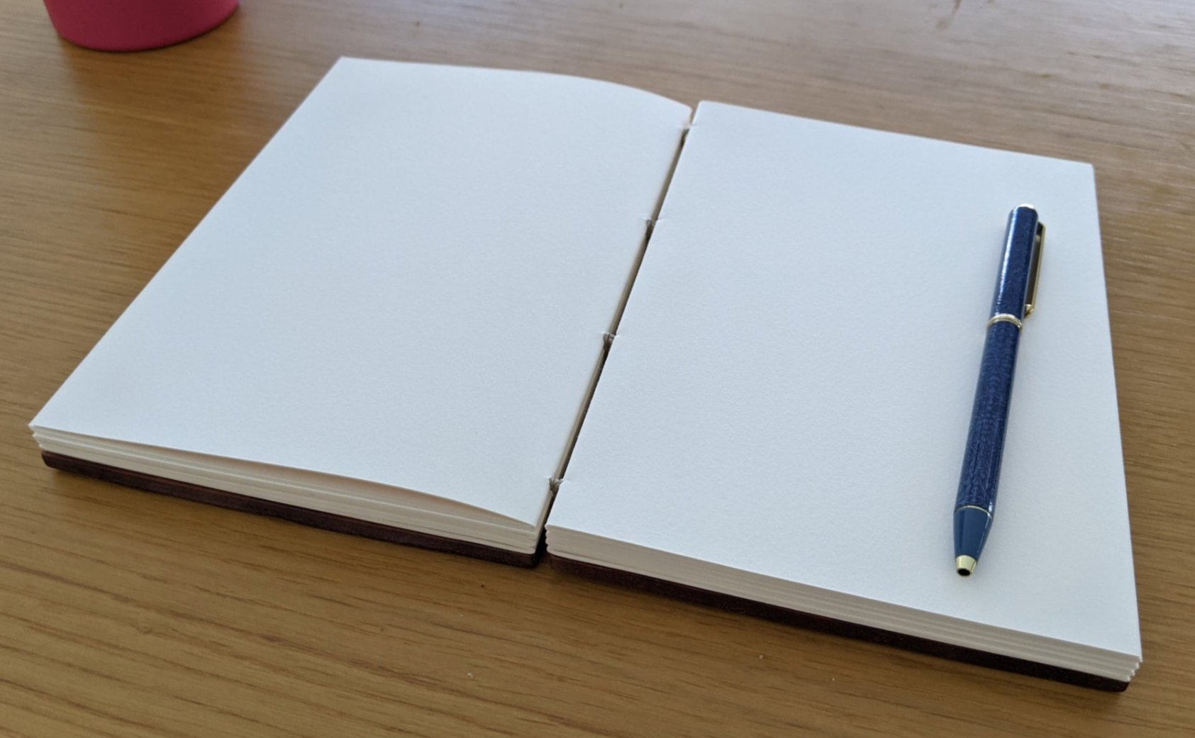 An open handcrafted wooden Coptic journal showcasing high-quality blank pages, ideal for writing, sketching, or journaling. A sleek blue and gold pen rests on the right-hand side, emphasizing the journal’s lay-flat design for effortless creativity. This artisanal notebook is perfect for artists, writers, and journal enthusiasts seeking a durable, aesthetically pleasing, and functional writing companion.