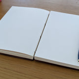 An open handcrafted wooden Coptic journal showcasing high-quality blank pages, ideal for writing, sketching, or journaling. A sleek blue and gold pen rests on the right-hand side, emphasizing the journal’s lay-flat design for effortless creativity. This artisanal notebook is perfect for artists, writers, and journal enthusiasts seeking a durable, aesthetically pleasing, and functional writing companion.