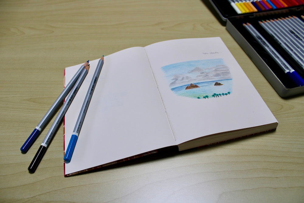 An open sketchbook lies on a wooden table, featuring a colored pencil drawing of two small islands with palm trees in the foreground and a cloudy sky above. Three colored pencils are placed across the left page, and a box of various colored pencils is visible in the background. The sketchbook captures the tranquil scene of the islands, showcasing an artistic moment and the tools used for creating the artwork. The overall setting is calm and conducive to creativity.